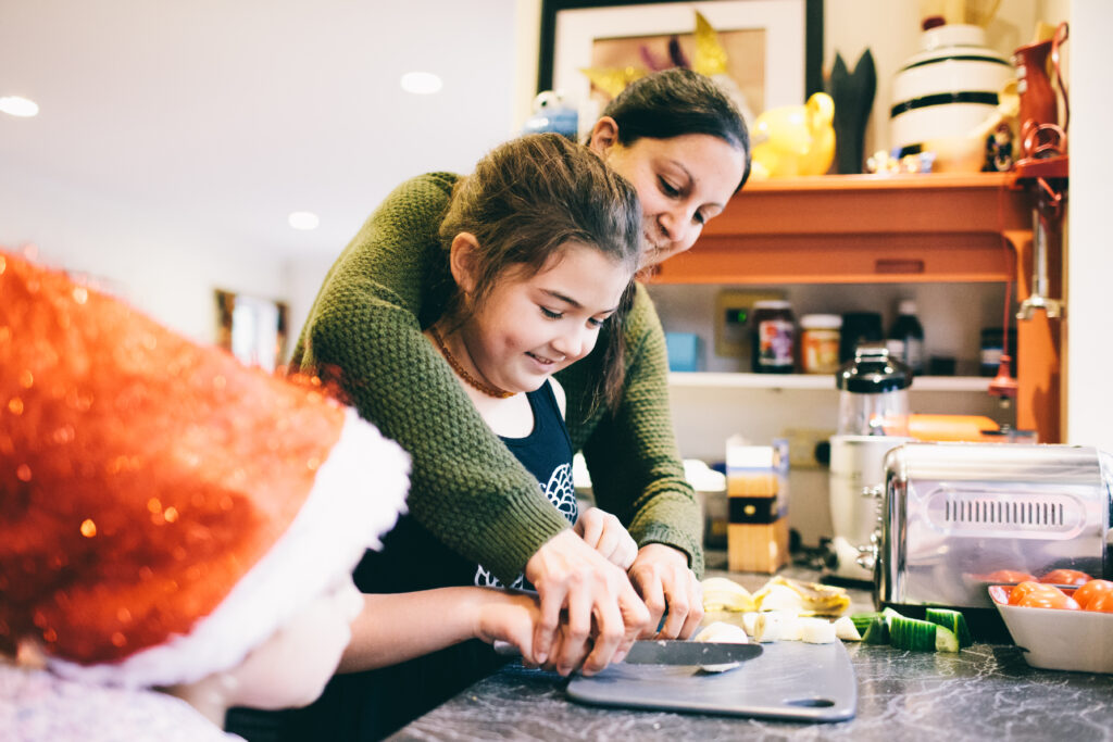 Blind Low Vision NZ. A mum and daughter in the kitchen chopping food. A young child looks on.